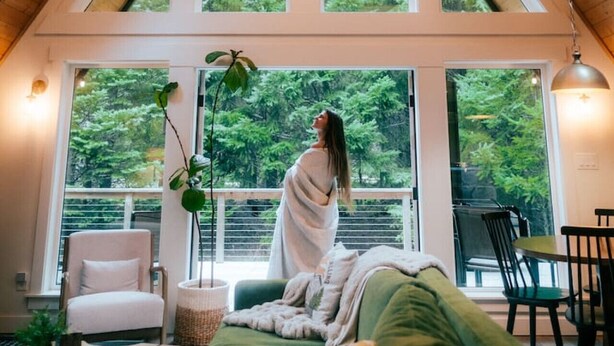 Una habitación con techo inclinado, ventanas amplias, cama, silla, mesa y una planta.