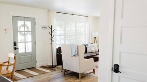 Quarto com piso de madeira, porta branca, cabideiro, sofá, cadeira e janela com cortinas.