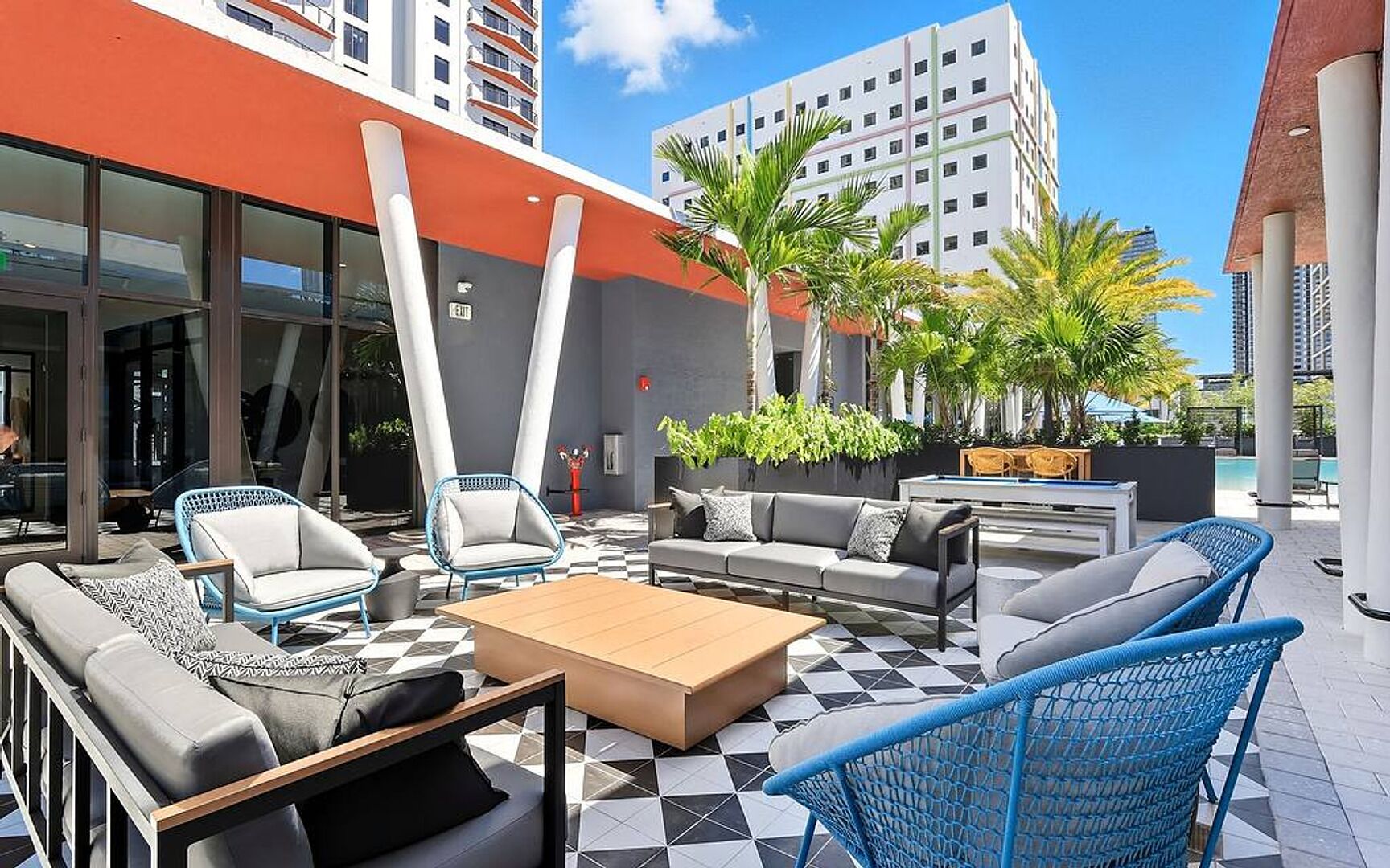 An outdoor seating area with sofas, chairs, and a coffee table, surrounded by palm trees and a pool.