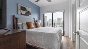 Een moderne slaapkamer met een bed, nachtkastje, lamp en stadszicht door een groot raam.