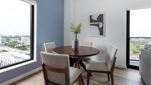 Een eetgedeelte met een ronde tafel, vier stoelen, een vaas met een plant en een stadszicht door grote ramen.