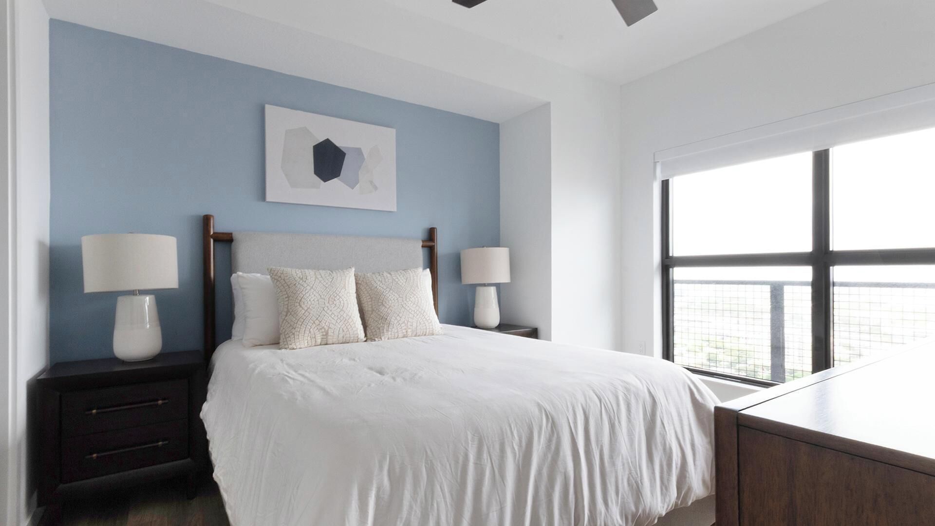 Une chambre à coucher moderne avec un lit, deux tables de chevet, une commode, une fenêtre et une œuvre d’art accrochée au mur.