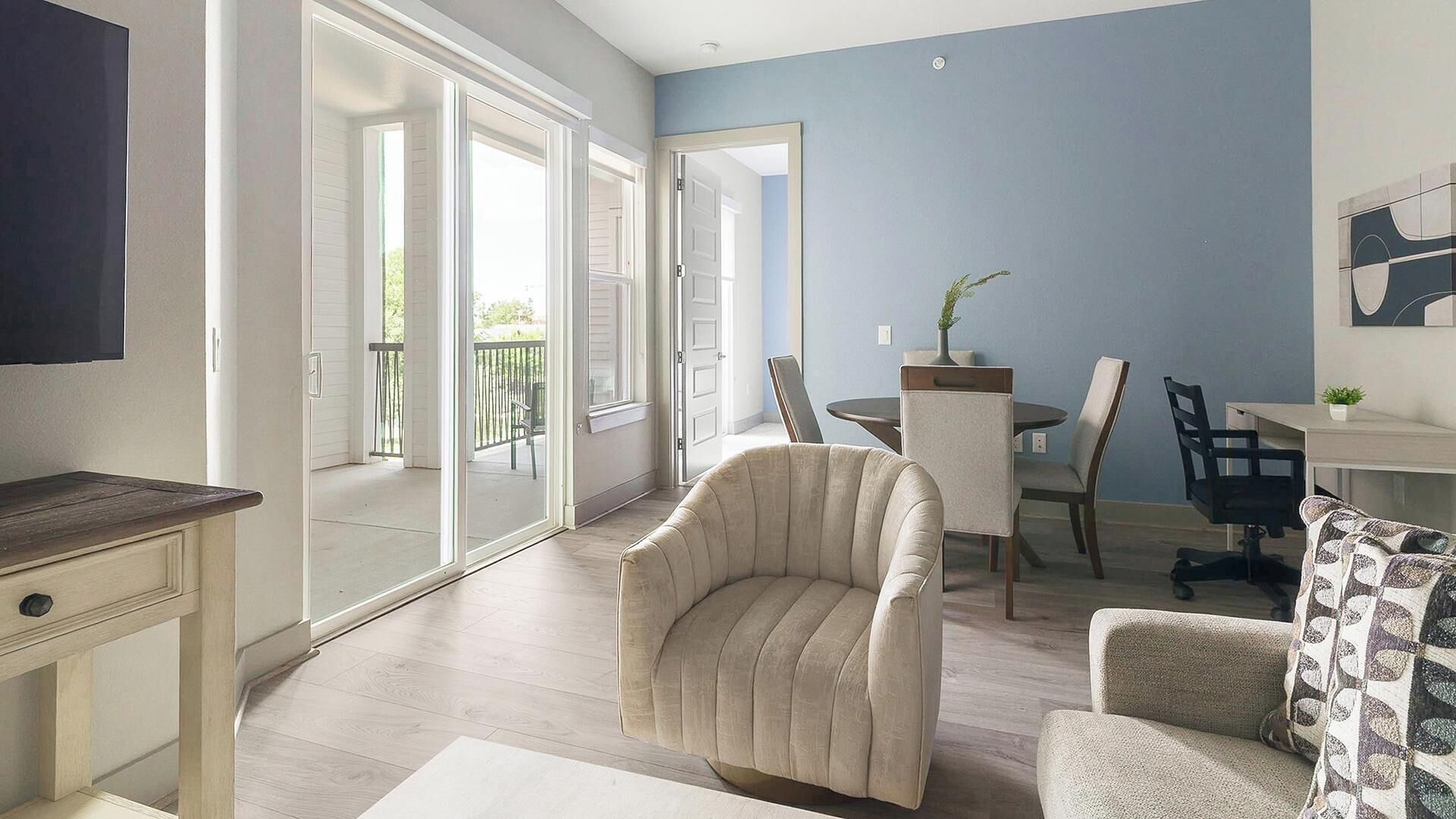 A room with a sliding glass door leading to a balcony, a dining table with chairs, a sofa, and a TV mounted on the wall.