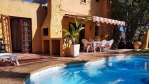 Una piscina amb terrassa enrajolada, zona de seients coberta a l'aire lliure i un edifici amb façana groga.