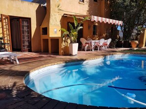 A swimming pool with a tiled deck, a covered outdoor seating area, and a building with a yellow facade. - Room-sunbird Room Double Room With poo (Cape Town)