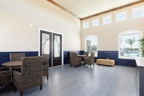 A spacious indoor area with a high ceiling, a seating arrangement with wicker chairs and a round table, and large windows providing natural light. - Landing | Beautiful 2BD, Clubhouse, Gym (Cocoa)