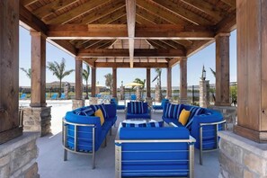 An outdoor seating area with blue sofas and tables, stone pillars, and a view of a pool and palm trees. - Landing | Beautiful 2BD, Clubhouse, Gym (Cocoa)