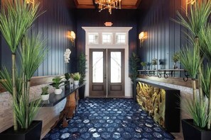 A hallway with dark wooden walls, a blue patterned floor, and a white fireplace. There are potted plants on either side and a bench with a metal frame. - Landing | Beautiful 2BD, Clubhouse, Gym (Cocoa)
