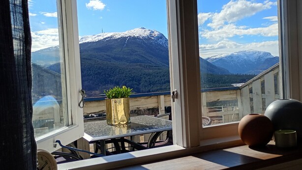A balcony with a view of snow-capped mountains, a potted plant, and a table with a clock.