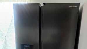 A Samsung refrigerator with a digital display, positioned in a room with wooden flooring and a window with lace curtains.