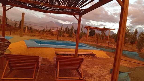 Une structure en bois formant une pergola, avec un espace piscine, entourée d’herbes sèches et d’arbres.