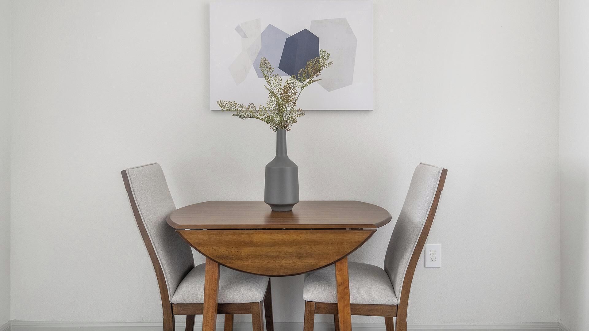 A round dining table with two chairs, a vase with flowers, and a piece of abstract art on the wall.