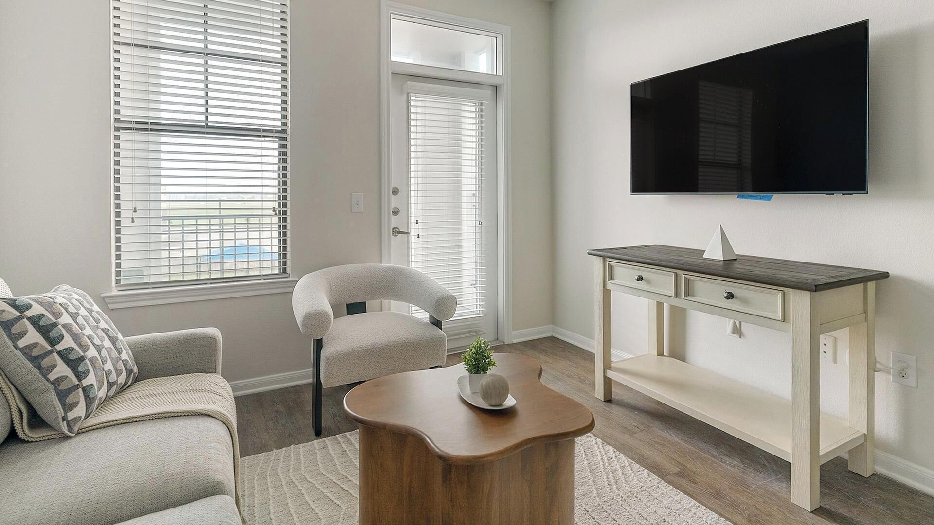 Een woonkamer met een bank, een fauteuil, een salontafel, een aan de muur bevestigde televisie en een raam met zonwering.