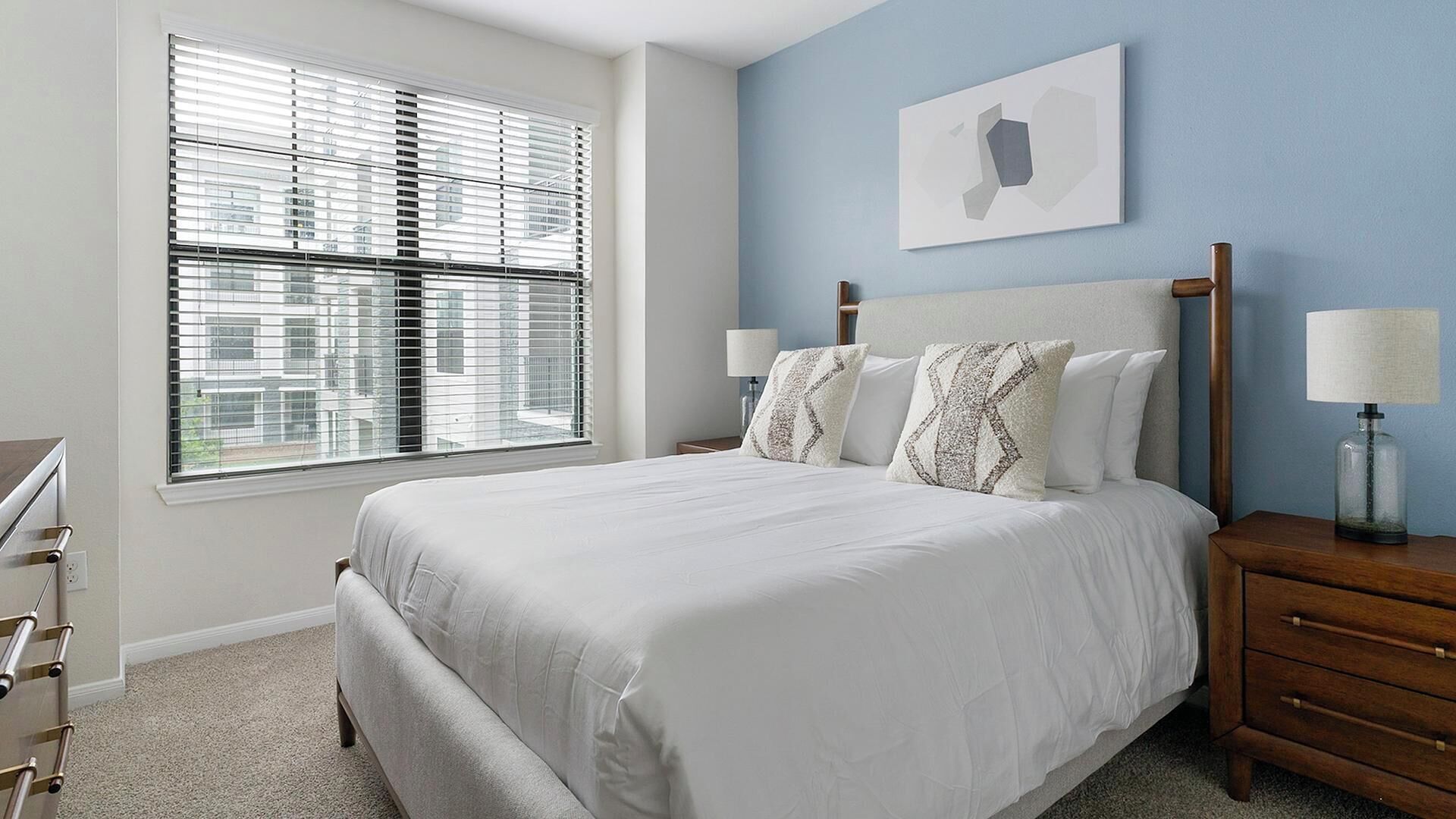 A bedroom with a large bed, a nightstand with a lamp, a dresser, and a window with blinds.