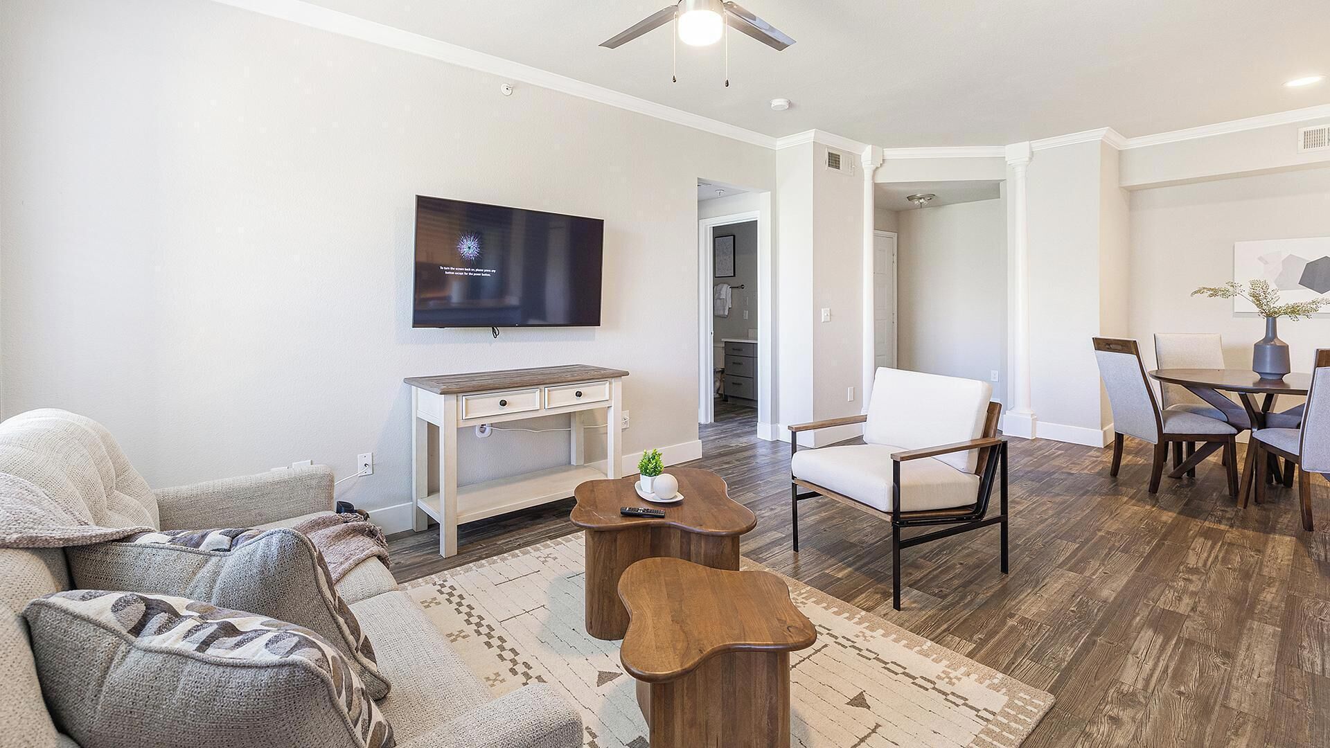 A modern living room with a flat-screen TV, a wooden desk, a white armchair, and a dining area with a table and chairs.