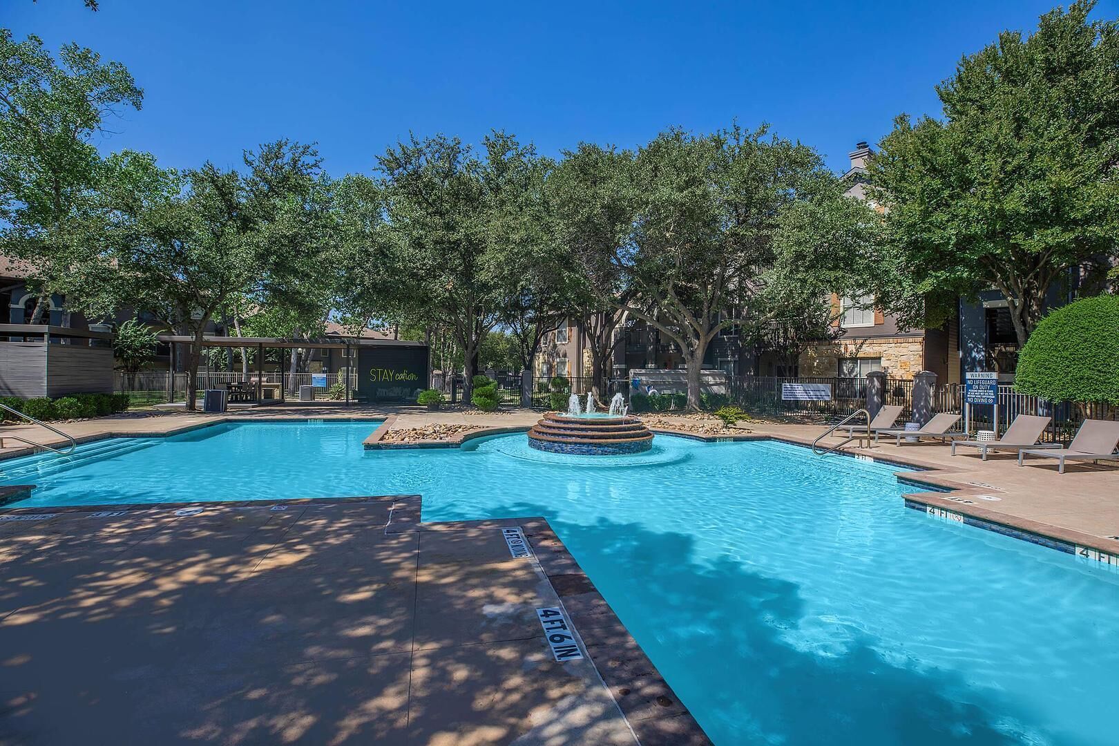 A swimming pool with a fountain, surrounded by trees and a covered seating area.