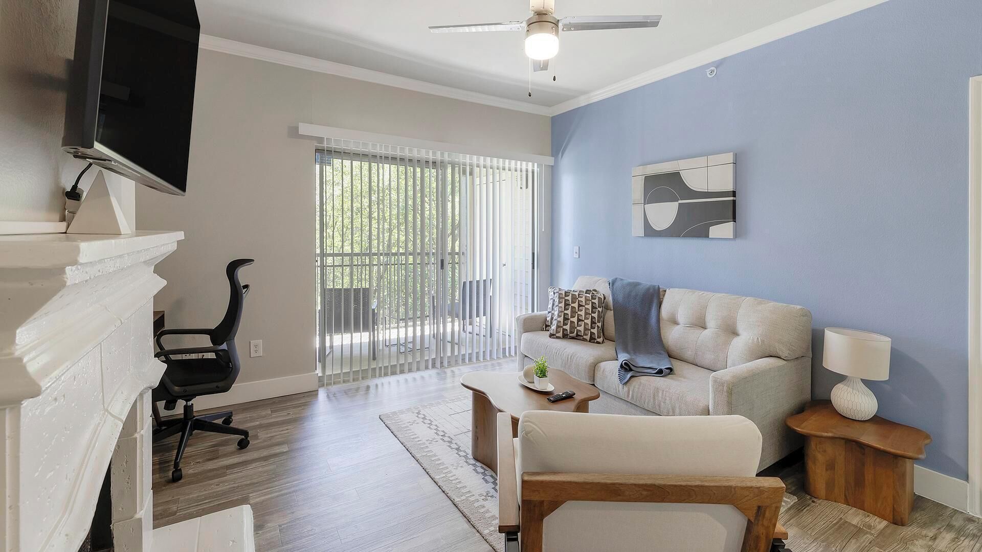 Un salon avec une cheminée, un téléviseur, un ventilateur de plafond, un canapé, une table basse et une vue sur l’extérieur à travers une fenêtre munie de stores.