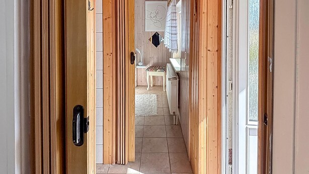 Un pasillo con puertas de madera, piso de baldosas y vista a una habitación con lavamanos y una silla.