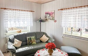 Una habitación con un sofá verde, una mesita redonda con un florero y dos ventanas con cortinas.