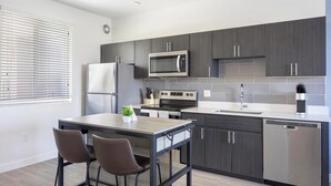 Una cocina moderna con muebles oscuros, electrodomésticos de acero inoxidable, una zona de comedor con dos sillas y una ventana con persianas.