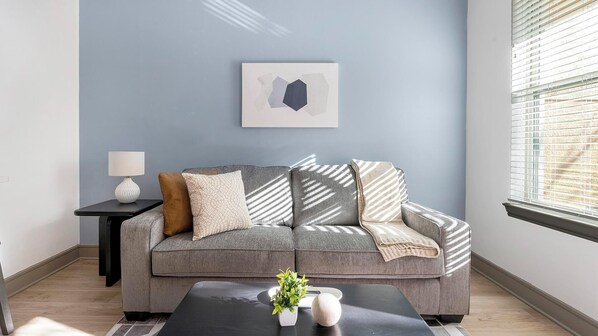 Ein Wohnzimmer mit grauem Sofa, einem kleinen Beistelltisch mit Lampe, einem gerahmten abstrakten Kunstwerk und einem Fenster mit Jalousien.