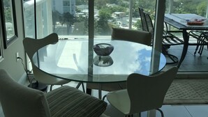 Ein modernes Wohnzimmer mit Glastisch, zwei Sesseln, einer Couch und Blick auf eine Stadtkulisse durch große Fenster.