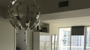 Ein modernes Wohnzimmer mit Glastisch, einer Couch, einem Essbereich mit Stühlen, einem weißen Schrank und einem großen Fenster mit Blick auf Gebäude.
