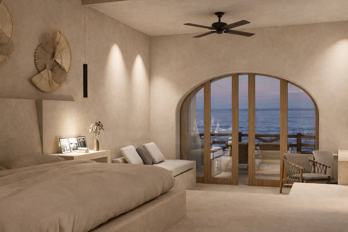 A bedroom with a large bed, a desk, a chair, a view of the ocean through a glass door, and a ceiling fan.
