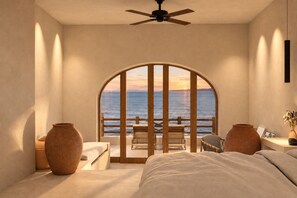 A bedroom with a large arched window overlooking the sea, a bed, two side tables, and a ceiling fan.