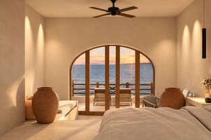 Un dormitorio con un gran ventanal arqueado con vistas al mar, una cama, dos mesitas de noche y un ventilador de techo.