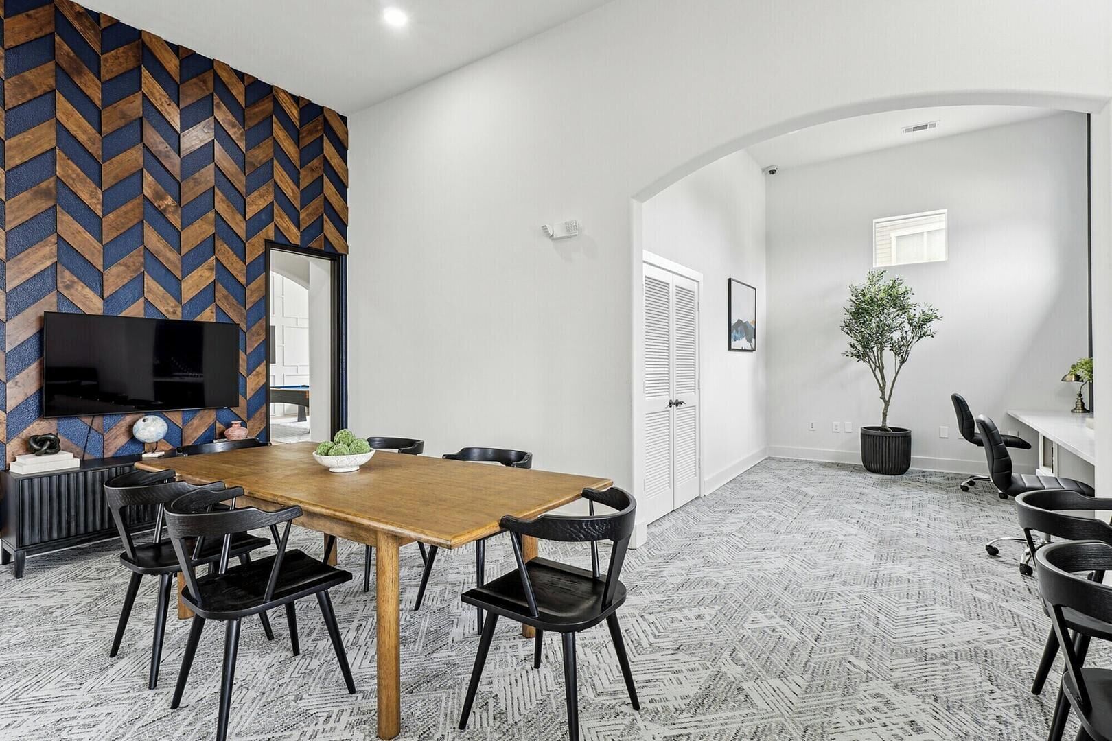 Een moderne vergaderruimte met een houten tafel, zwarte stoelen, een groot flatscreen-televisie en een kamerbreed tapijt met dessin.
