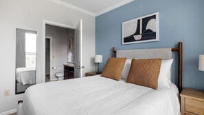 A hotel room with a large bed, a nightstand, a wall-mounted lamp, and a bathroom visible through an open door. - Landing | Amazing 2BD, Clubhouse, Gym (Houston)