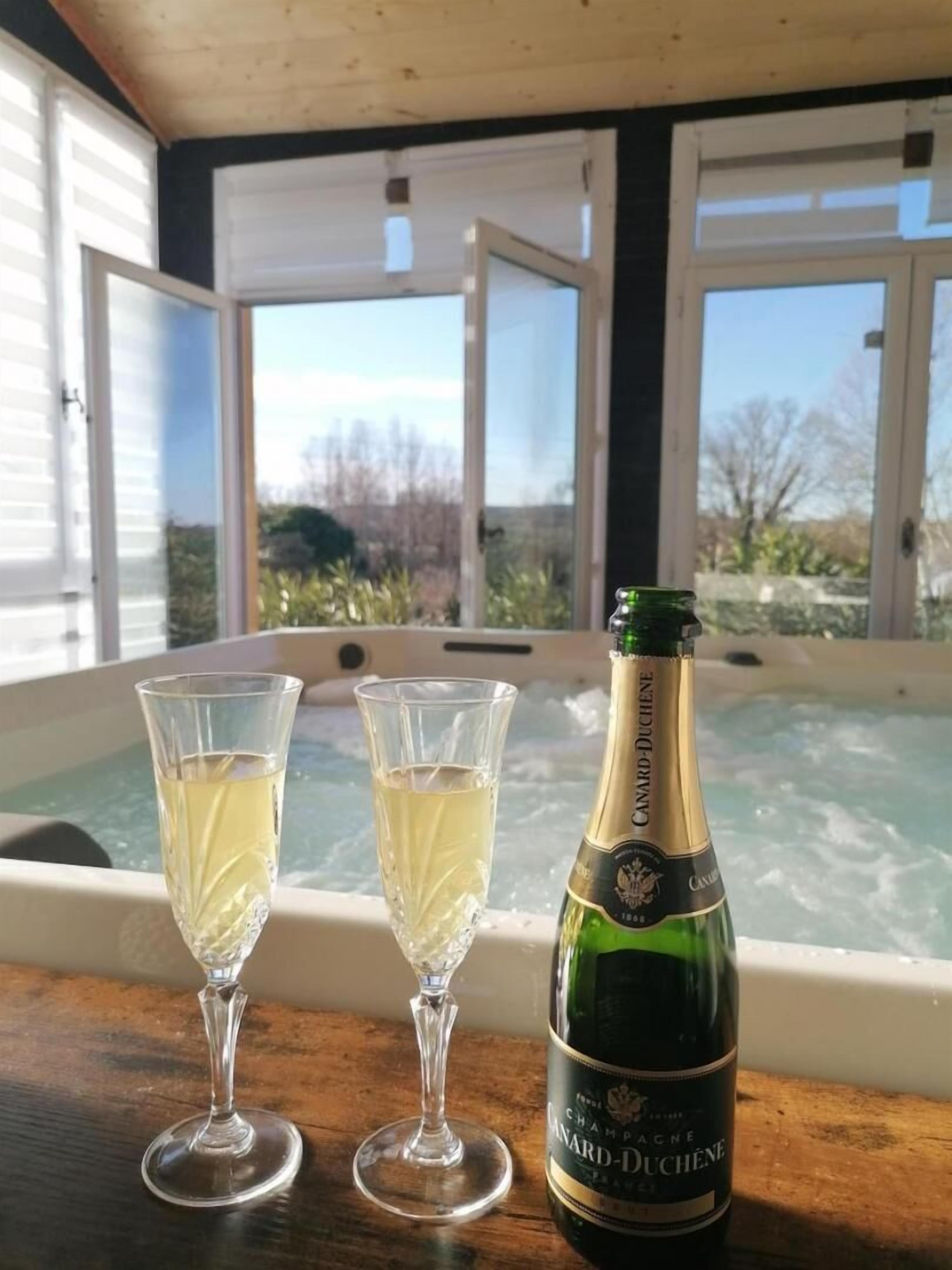 Two champagne flutes and a bottle of champagne on a wooden table with a hot tub in the background.