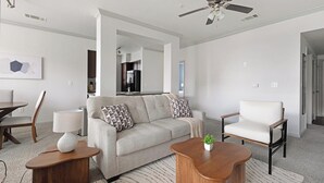 A modern living room with a sofa, a chair, a coffee table, and a lamp. There is a ceiling fan and a kitchen visible in the background. - Landing | Stylish 2BD, Game Room, Clubhouse (Houston)