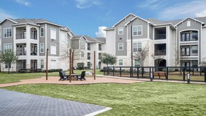 A residential complex with multiple-story buildings, outdoor seating, and a paved walkway. - Landing | Amazing 2BD, Clubhouse, Game Room (Houston)