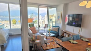 A modern living room with a large window, a flat-screen TV, a dining table, and a sofa. - Mountain View l Gym, Patio & Parking, 5 mins to DT (Vancouver)