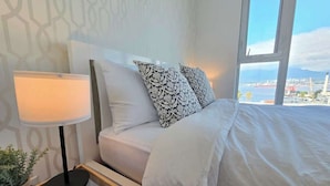A hotel room with a bed, a nightstand, a lamp, and a view of the city. - Mountain View l Gym, Patio & Parking, 5 mins to DT (Vancouver)