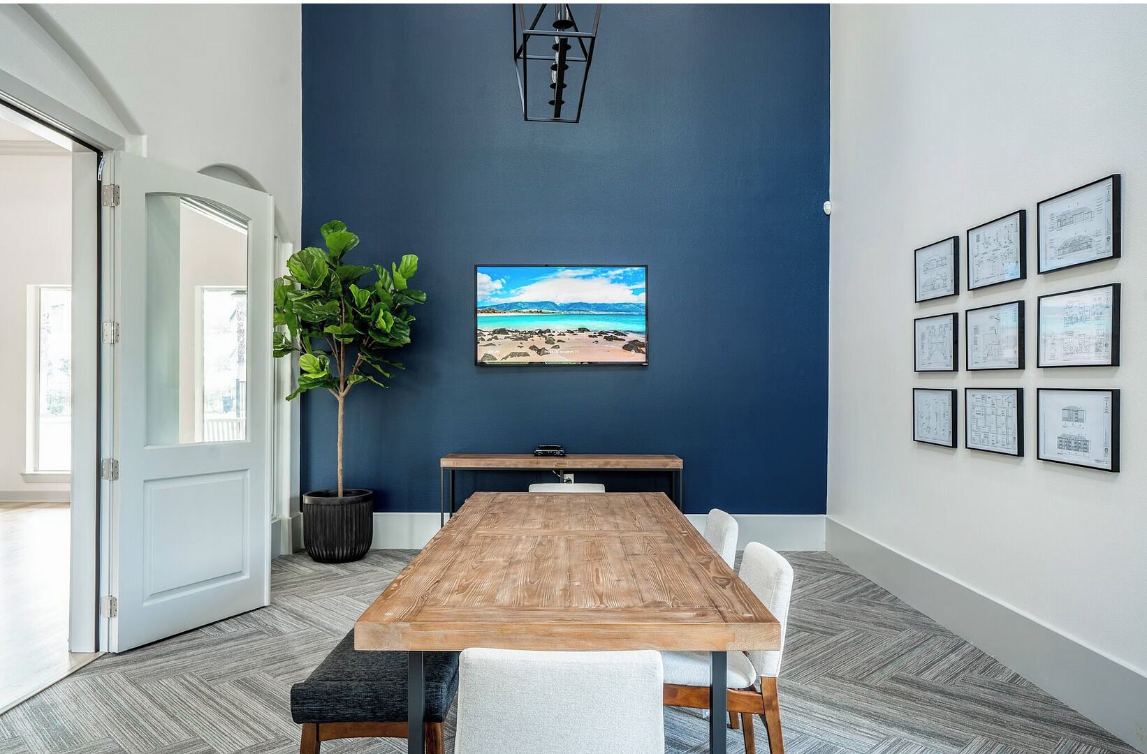 A conference room with a wooden table, white chairs, a potted plant, a framed picture of a beach, and architectural drawings on the wall.