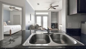 A modern kitchen with a double sink, a dining area with a table and chairs, a sofa, and a ceiling fan. - Landing | Chic 1BD, Clubhouse, Game Room (Houston)