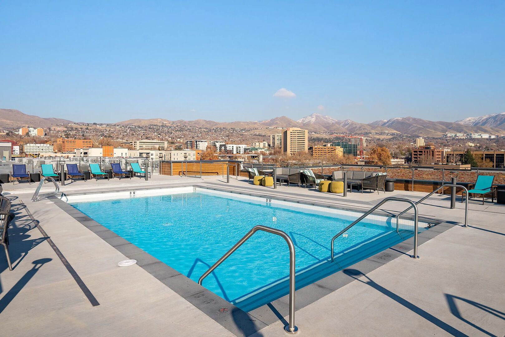 A rooftop pool with clear blue water, surrounded by lounge chairs and a view of mountains and buildings in the background.