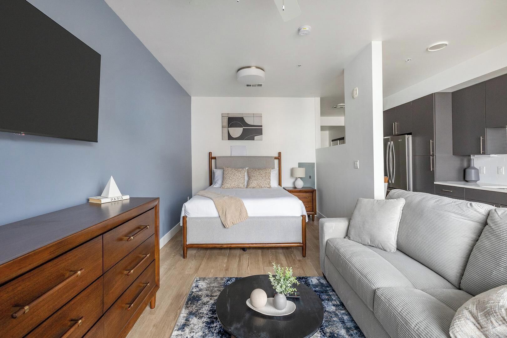 A modern hotel room with a bed, a sofa, a wooden dresser, a flat-screen TV, and a kitchenette.