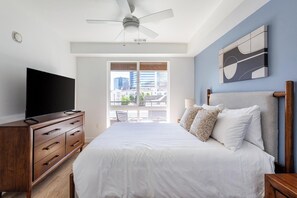 Quarto com cama, penteadeira, televisão, ventoinha no teto e vista para edifícios através de uma janela.