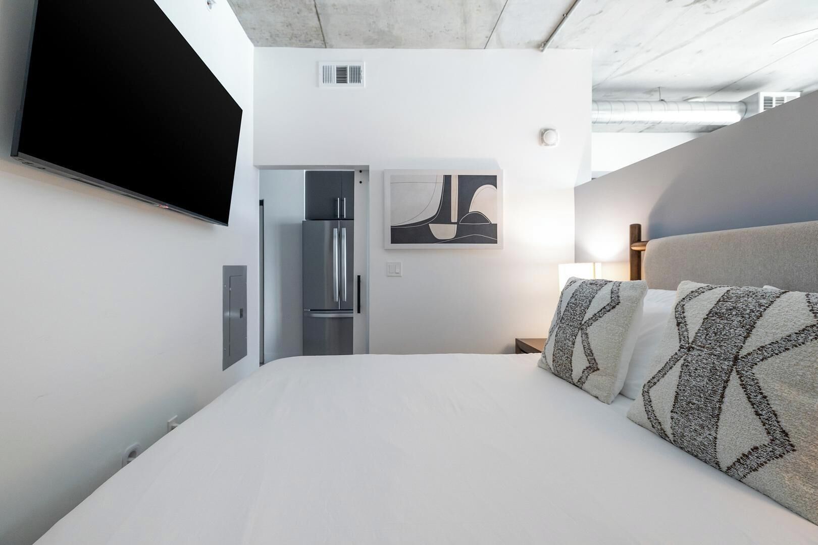 A modern hotel room with a bed, a flat-screen TV, a refrigerator, and a painting on the wall.