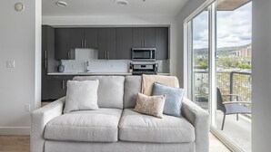 En moderne stue med en grå sofa, en balkon med udsigt og et køkken med mørke skabe og en mikrobølgeovn.