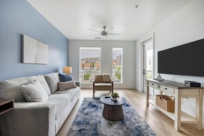 A modern living room with a sofa, a coffee table, a TV, and a view of buildings through large windows. - Landing | Amazing 1BD in Central City (Salt Lake City)