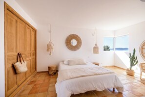 Une chambre avec un lit, une armoire en bois, une suspension en macramé, un miroir rond et une vue sur l’extérieur.