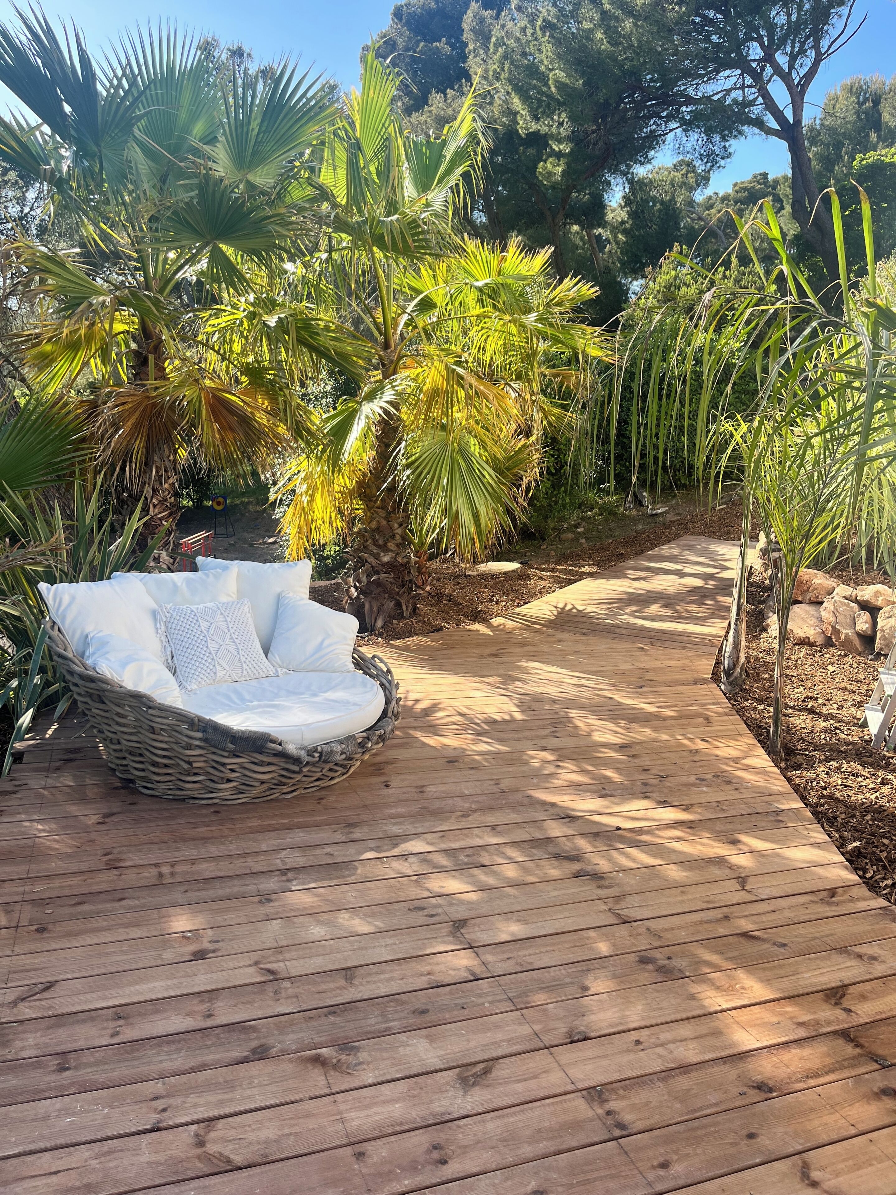 Eine Holzterrasse mit einem Korbstuhl und Kissen, umgeben von tropischen Pflanzen und Bäumen.