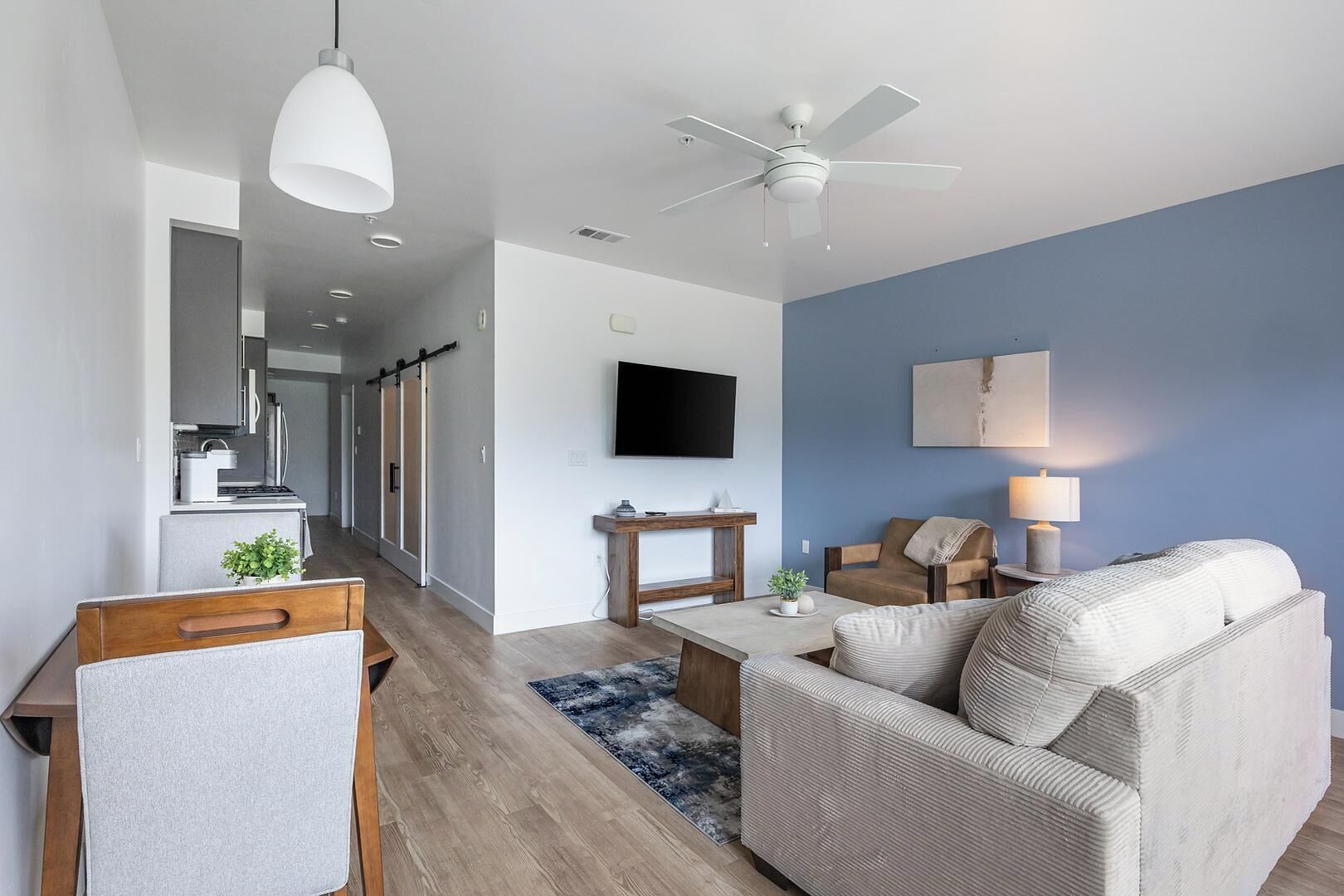 A modern living room with a sofa, coffee table, and TV. There is a kitchen area with cabinets and a ceiling fan.