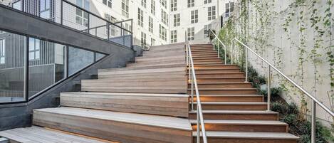 A staircase with wooden steps and metal railings, leading to a modern building with large windows and a concrete wall with climbing plants.