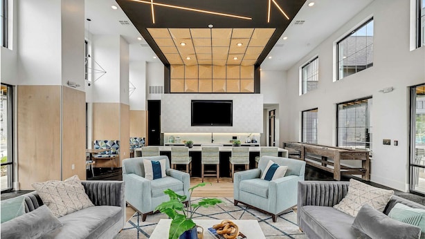 A modern living room with grey sofas, a coffee table with a plant and a bowl of pretzels, and a kitchen area with a TV above the counter.